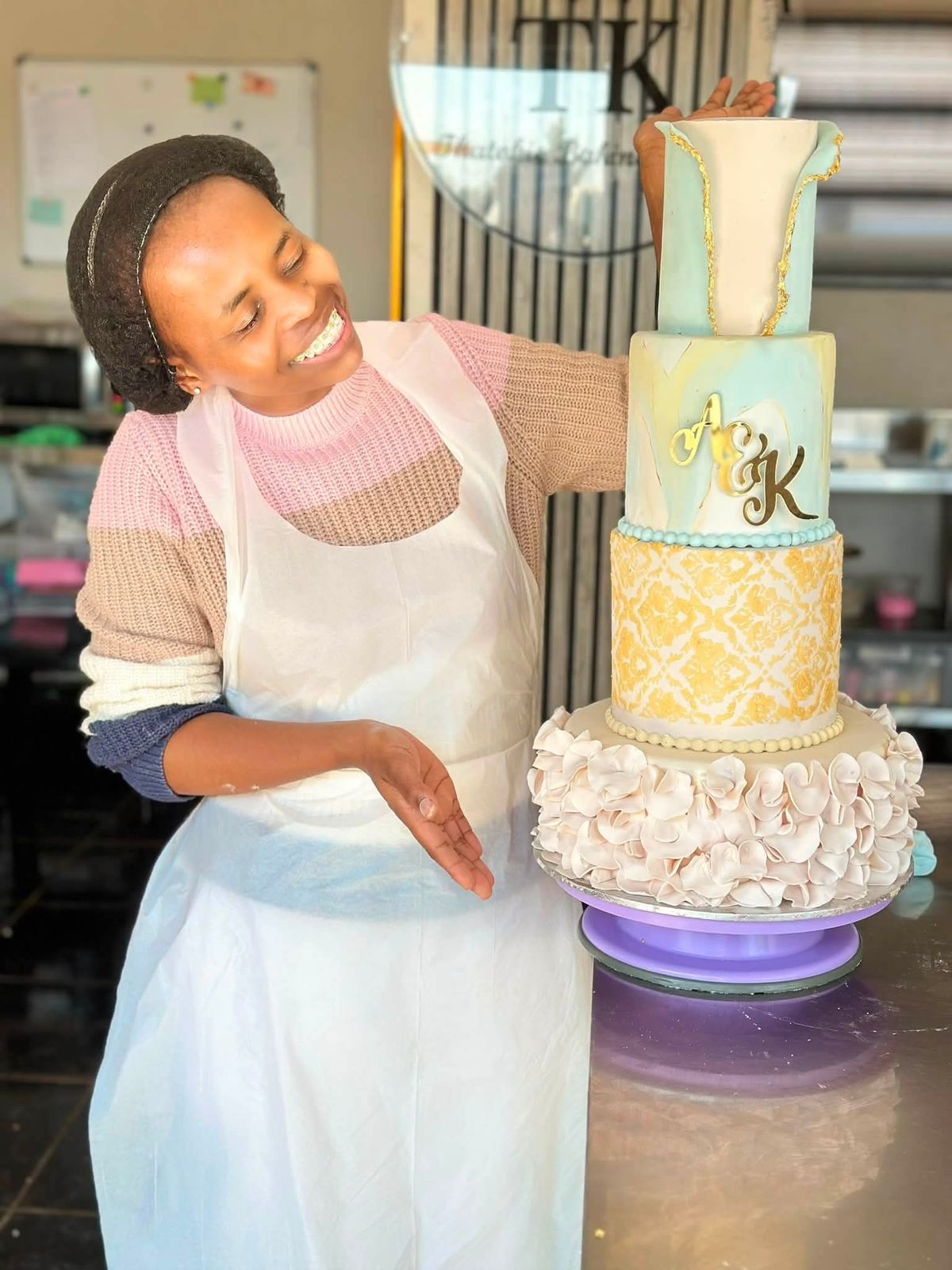 Lerato smiling beside a four-tier wedding cake with sugar ruffles, gold damask and gold-leaf edges
